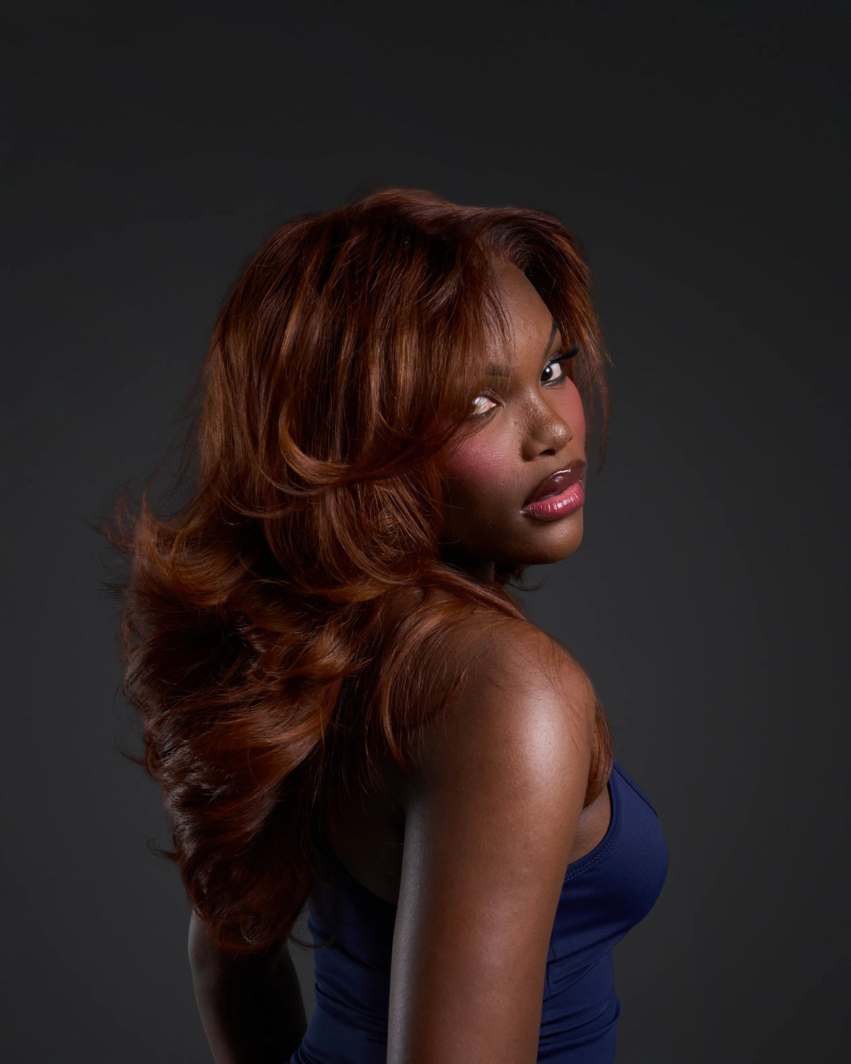A woman with long, wavy auburn hair wearing a dark blue strapless top, photographed against a dark gray studio background.