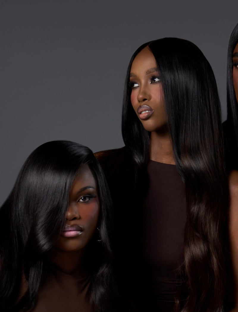 Three women with long, straight, dark brown to black hair pose together in a studio setting against a dark gray background. They are wearing matching dark brown sleeveless dresses and gold jewelry.
