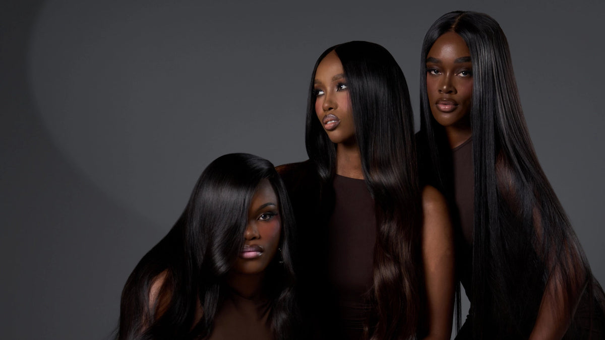 Three women with long, straight, dark brown to black hair pose together in a studio setting against a dark gray background. They are wearing matching dark brown sleeveless dresses and gold jewelry.