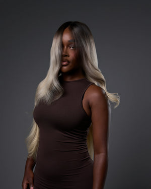 A woman modeling a long, wavy hair extension or wig with a dark brown root and blonde ombré ends, wearing a dark brown sleeveless dress against a dark gray studio background.